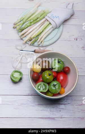 Weißer und grüner Spargel auf einem Holzbrett, Spargelschälungen, ein Schäler und eine Schüssel mit verschiedenen Tomaten Stockfoto