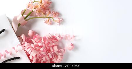 Rosa Frühlingsblumen, rosa Geschenkstreamer in weißer Geschenktüte auf weißem Hintergrund. Wunderschöne Frühlingskarte für Geburtstag, Frau, Valentinstag, Muttertag in Pastellfarben. Horizontale, flache Verlegung. Speicherplatz kopieren Stockfoto