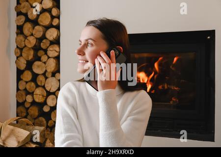 Lächelnde junge kaukasische Frau in weißem Pullover, die auf warmem Boden in der Nähe des Kamins zu Hause sitzt und sich mit dem Handy unterhält Stockfoto