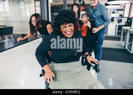Spaß mit den Kollegen haben. Glückliche Geschäftsfrau lacht fröhlich, während sie im Büro auf einen Stuhl geschoben wird. Geschäftsfrau genießt Spielzeit mit Stockfoto