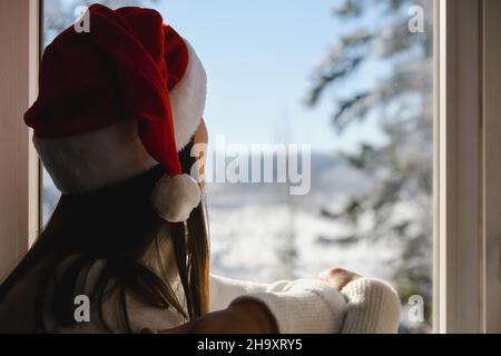 Die selektive Konzentration der jungen Frau in stilvollem weißen Strickpullover und Santa Red Hut sitzt zu Hause auf der Fensterbank sieht den schönen verschneiten Wald Stockfoto