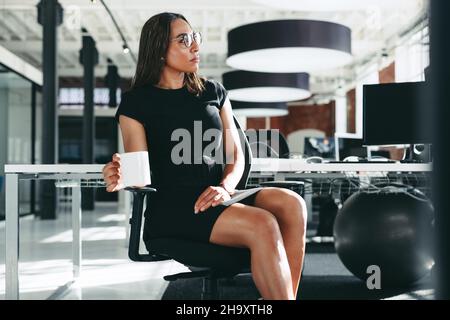 Durchdachte Geschäftsfrau, die allein in einem Büro sitzt. Selbstbewusste junge Geschäftsfrau schaut weg, während sie auf einem Stuhl mit einer Tasse Kaffee in ihrem h sitzt Stockfoto