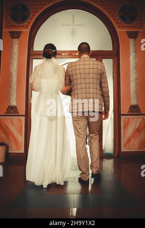 Um 2021: Das Brautpaar hält die Hände in der Kirche. Blick von hinten. Hochzeitstag Konzept.schöne Kirche Bogen und Wände. Hochwertige Fotos Stockfoto