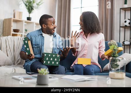 Überglücklich sitzt ein junges Paar auf der Couch mit bunten Geschenkschachteln in den Händen. Afrikanischer Mann und Frau öffnen ihre Geschenke während der Feiertage. Aufrichtige Emotionen während unerwarteter Überraschung. Stockfoto