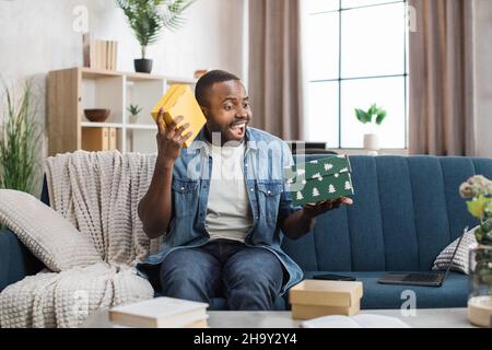 Aufgeregter afrikanischer junger Mann im Denim-Hemd, der bunte Geschenkschachteln öffnet, während er auf der bequemen Couch sitzt. Konzept von Menschen, Urlaub und Überraschungen. Stockfoto