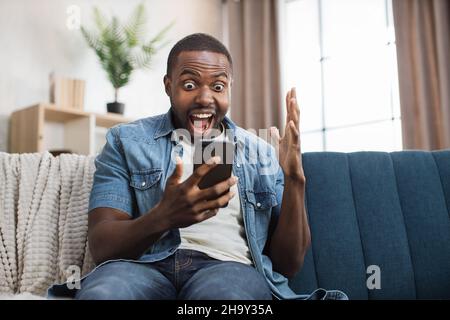 Überglücklich, afroamerikanischer Mann, der Erfolg mit Ja-Geste feiert, während er einige Nachrichten über Waren auf dem Handy erhält. Junger Mann, der mit aufrichtigem Glück im Gesicht auf der Couch sitzt. Stockfoto
