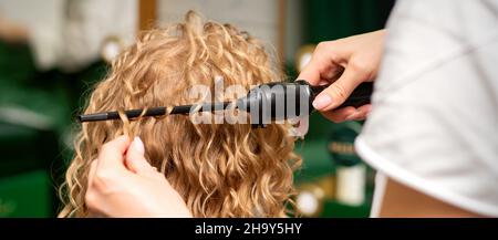 Hairstylistin-Hände lockern welliges Haar einer jungen Frau, die ein Lockenstab für Haarlocken in der Rückansicht des Schönheitssalons verwendet Stockfoto