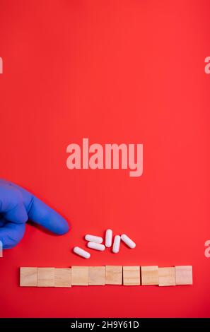 Hände in medizinischen Handschuhen zeigen auf Holzklötze auf farbigem rotem Hintergrund mit Medikamentententabletten. Konzeptbuch über medizinisch-wissenschaftliche oder biologische Temp Stockfoto
