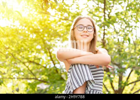 Porträt eines lächelnden Mädchens, das eine Brille trägt und die Arme auf einem sonnigen bewaldeten Hintergrund über ihr Knie gekreuzt sind Stockfoto