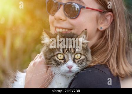 Nahaufnahme eines lächelnden Mädchens mit Sonnenbrille, das eine niedliche Katze in den Armen hält und auf die Kamera schaut Stockfoto