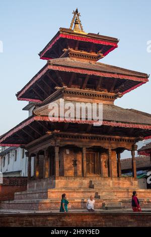 Am frühen Morgen sitzt ein Mann auf dem Sockel des Mahabishnu-Tempels, während Frauen am Durbar Square, Kathamandu, Nepal, vorbei gehen. Stockfoto