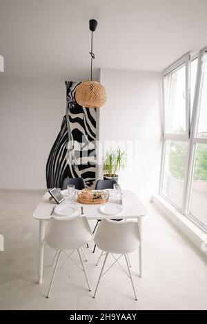 Innenarchitektur eines hellen Speisesales in einem minimalistischen Stil mit einem quadratischen weißen Holztisch, schwarzen und weißen Stühlen, mit Korbdekorationen auf dem Kronleuchter und auf dem Tisch, mit einem großen Fenster und einem riesigen Zebra-Bild an der Wand. Hochwertige Fotos Stockfoto