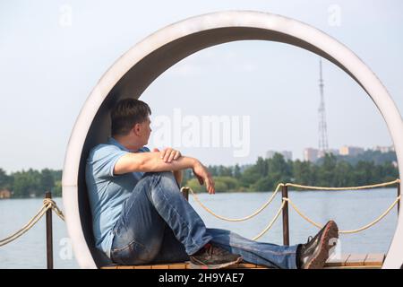 Ein erwachsener Mann sitzt in Form eines Kreises auf einer kreativen Bank und blickt auf die ruhige Oberfläche des Flusses. Stockfoto