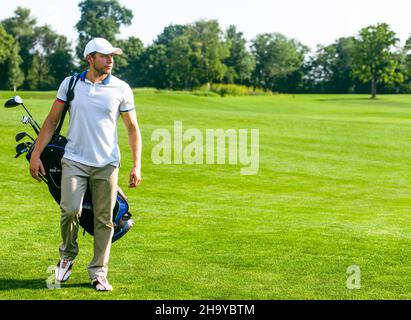 Der junge Golfer trägt eine Tasche mit Golfschlägern. Schlanker Mann in weißem T-Shirt, Golfschuhen, weißer Mütze und beigefarbener Hose geht in das grüne Turf Grass. Speicherplatz Kopieren. In voller Länge. Hochwertige Fotos Stockfoto