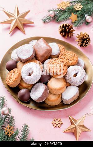 Weihnachtsdelikatessen Polvorones und Mantecados in goldenem Teller auf rosafarbenem Tisch, verziert mit goldenen Kugeln, christbaumzweigen und Tannenzapfen, oben Stockfoto