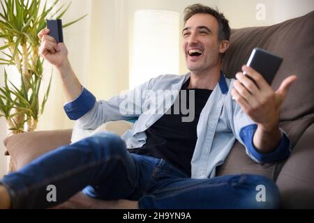 Glücklicher Mann, der online von einem Smartphone kauft, das zu Hause auf dem Sofa liegt. Horizontale Zusammensetzung. Stockfoto
