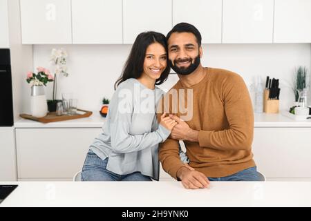 Süßes multiethnisches Paar in der Liebe, das in Umarmungen in der Küche steht. Fröhliche Frau und ihr Mann schauten auf die Kamera und lächelten toothy Stockfoto