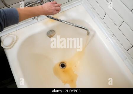 Weibliche Hand öffnet einen Wasserhahn, aus dem verrosttes Wasser in ein weißes Bad fließt. Das Problem sind alte Rohre, die ausgetauscht werden müssen. Stockfoto