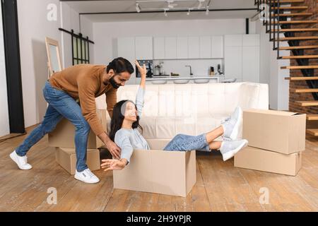 Glückliches multirassisches Paar zum ersten Mal zu Hause Käufer Spaß beim Auspacken Habseligkeiten am bewegenden Tag. Aufgeregt Frau Reiten in Karton, während ihr Mann schieben es in neue Hauswohnung Stockfoto