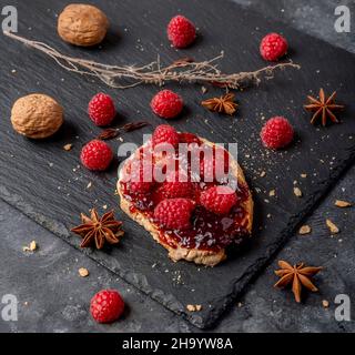 Ein köstlicher Crouton mit Marmelade und ganzen Himbeeren oben und ein paar Walnüssen daneben, auf schwarzem Schiefergrund Stockfoto