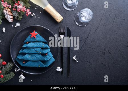 Weihnachtliche Tischausstattung in schwarzer Farbe mit schwarzem Besteck, Serviette als Weihnachtsbaum gefaltet, weihnachtliche Silberkugeln, Champagner, Gläser auf schwarzem Rücken Stockfoto