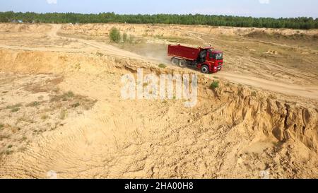 Muldenkipper fährt auf Landstraße. Szene. Draufsicht auf LKW-Fahrten, die vor dem Hintergrund der Baggerkarriere Staubwolken auf unbefestigten Straßen auf dem Land hinterlassen Stockfoto