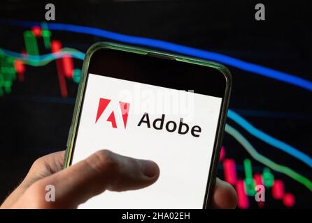 In dieser Fototafel ist das amerikanische multinationale Computer-Multimedia- und Kreativitäts-Software-Unternehmen Adobe-Logo auf einem Smartphone mit einem wirtschaftlichen Börsenindex-Diagramm im Hintergrund zu sehen. Stockfoto