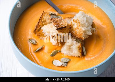 Cremige Butternut-Kürbissuppe mit Brottoast Croutons in blauer Keramikschale auf weißem Holzhintergrund, herbstlich gemütliches veganes Gericht Stockfoto