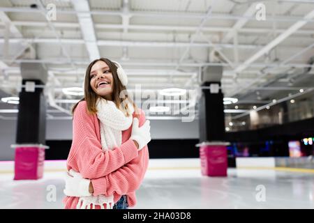 Erfreut Frau in Ohrenschützer und rosa Pullover umarmen sich auf Eisbahn Stockfoto