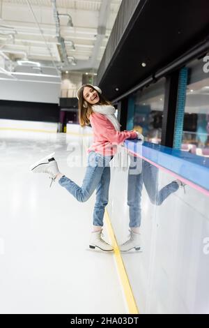Die ganze Länge der glücklichen Frau in Pullover, weißem Schal und Ohrenschützer posiert auf der Eisbahn Stockfoto