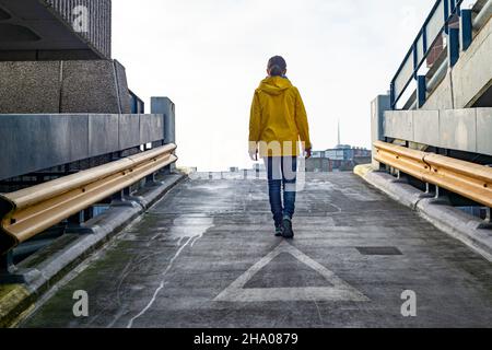 Rückansicht einer Frau, die im Regen eine Parkrampe hochgeht und einen gelben Mantel trägt. Stockfoto