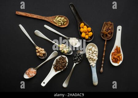 Set von unordentlichen Löffeln aus verschiedenen Materialien und Formen, gefüllt mit Nüssen, Samen, Gewürzen und Hülsenfrüchten auf schwarzem Hintergrund Stockfoto