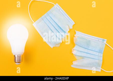 Leuchtende Energiesparlampe mit zerrissener medizinischer Gesichtsmaske auf gelbem Hintergrund - Anti-Maskenkonzept Stockfoto