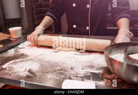 Eine Bäckerin trägt eine Schürze, während sie Teig auf dem Küchentisch ausrollt Gebäck Backkonzept Kuchen backen Kekse backen hausgemachte Pizza in einem Stockfoto