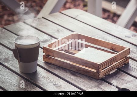 Einzelne Papier Take away Tasse Kaffee mit heißem Dampf auf dem Holztisch hinter einer Box mit Servietten Stockfoto