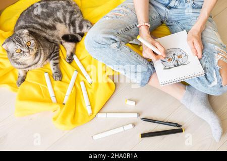 Zeichnen zu Hause. Ein Mädchen in blauen Jeans skizziert eine graue Falte Katze mit gelben Augen. In der Nähe sind Marker, ein gelbes Karli. Die Katze sitzt in der Nähe des Mädchens. V Stockfoto