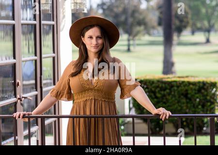 Frau in einem langen braunen Kleid mit einem Hüfthut und Händen am Geländer Stockfoto