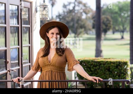 Lächelnde Frau in braunem Kleid & einem Hipster-Hut mit Händen am Geländer Stockfoto