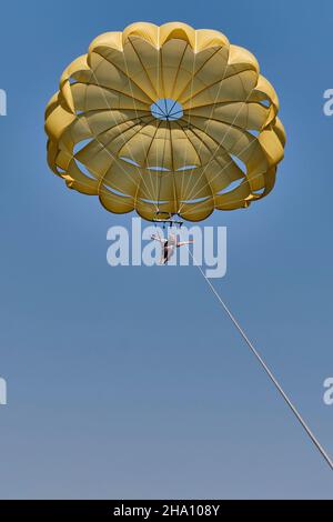 Eine ältere weiße Frau parasegelt.Sie fliegt unter einem gelben Fallschirm am blauen Himmel, lächelt und öffnet ihre Arme. Stockfoto
