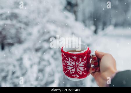 Nahaufnahme einer Tasse Tee oder Kaffee in rot gestricktem Weihnachtsmantel in weiblicher Hand im Winterpark. Getränke zum Aufwärmen bei schneebedecktem frostigen Wetter outdoo Stockfoto