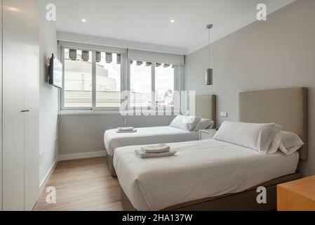 Schlafzimmer mit zwei separaten Betten mit Handtüchern und weißen Schränken, wandfernseher und großem Fenster mit Blick in eine Ferienwohnung Stockfoto