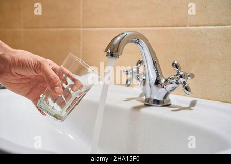 Mans Hand hält leeres Glas in der Nähe Badezimmer Edelstahl Säule Wasserhahn. Erwägen Sie, fließendes Wasser aus dem Chrom-Wasserhahn zu gießen. Sicher zu trinken oder nicht fit Stockfoto