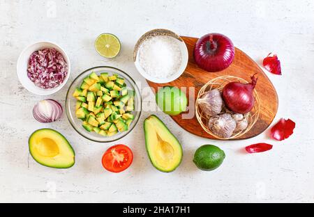 Avocado Hälften, Stücke, Limetten, Tomaten und Zwiebeln, Tasse mit Salz - Grundzutaten Guacamole auf weißem Arbeitsbrett, flach legen Foto. Stockfoto