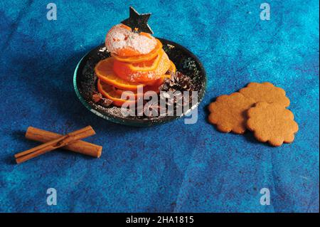 Aus orangefarbenen Scheiben gefertigte, winklige Ansicht eines winzigen Weihnachtsbaums. Blauer Backgroun mit Zimtstangen und Ingwerbrot. Stockfoto