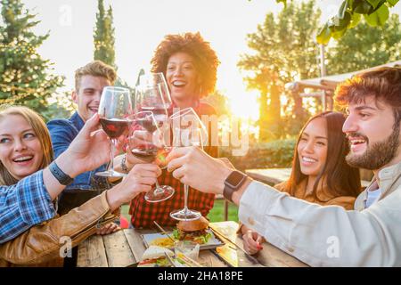 Gruppe multiethnischer Freunde, die einen gesunden Lebensstil leben und lächeln und scherzen, während sie Rotwein im Pub-Restaurant im Freien trinken - Junge Leute toasten Stockfoto