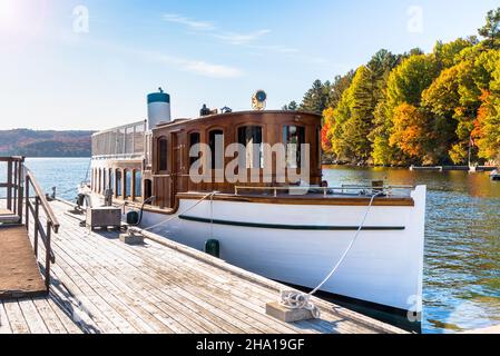 Altes Dampfschiff, das an einem sonnigen Tag an einem hölzernen Pier am See mit bunten Herbstbäumen im Hintergrund vertäut ist Stockfoto