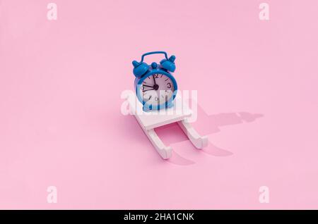Eine blaue Uhr, die zehn Minuten vor Mitternacht auf weißem Holzschlitten auf pastellrosa Hintergrund zeigt. Minimalistisches romantisches kreatives Konzept für Weihnachten oder Stockfoto