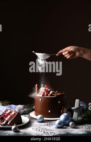 Frau, die auf dunklem Hintergrund leckere Weihnachtsschokoladenkuchen mit Puderzucker schmückt Stockfoto