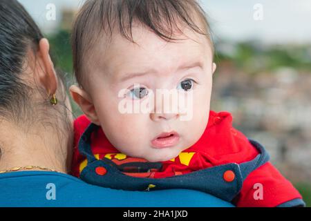 Schönes lateinisches Baby, das seinen Kopf über die Schulter seiner Mutter blickt, großes Baby mit neugierigem Blick, blauem Overall und rotem Hemd, das nach links schaut. Mit Hallo Stockfoto