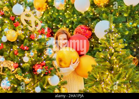 Ein fröhliches Mädchen hält drei große Weihnachtskugeln, die von funkelnden Weihnachtsbäumen umgeben sind. Neujahrsstimmung. Stockfoto